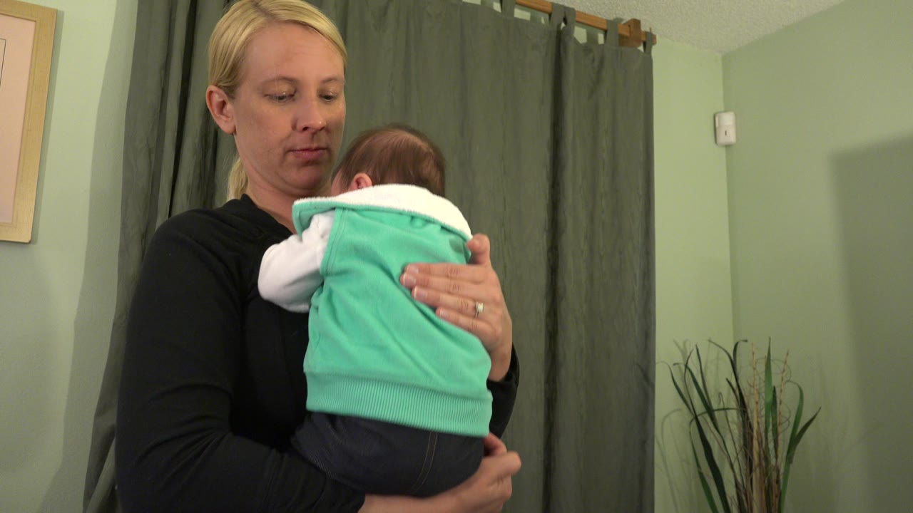 At 0:00, a blonde woman in black holds a baby in a turquoise vest close to her chest inside a Westminster, Colorado room with green walls and curtains.