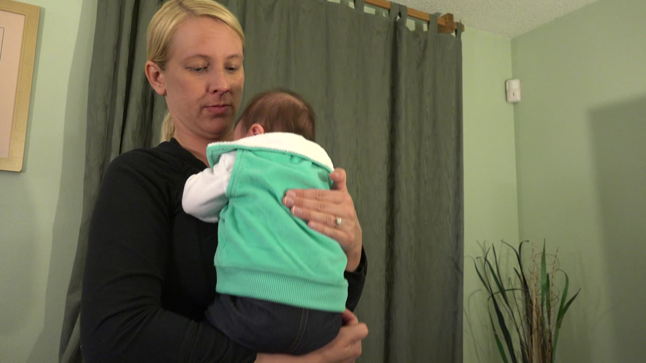At 0:01, the woman keeps the baby upright against her chest while green curtains and a small framed wall piece remain visible behind them in Westminster, Colorado.