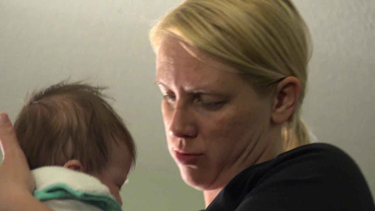 At 0:00 in Westminster, Colorado, a blonde woman in a black shirt holds a tiny infant partly turned away beside a plain pale wall.