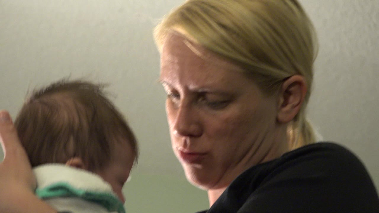 At 0:01 in Westminster, Colorado, the woman looks downward as the infant's head and white clothing remain close to her shoulder.
