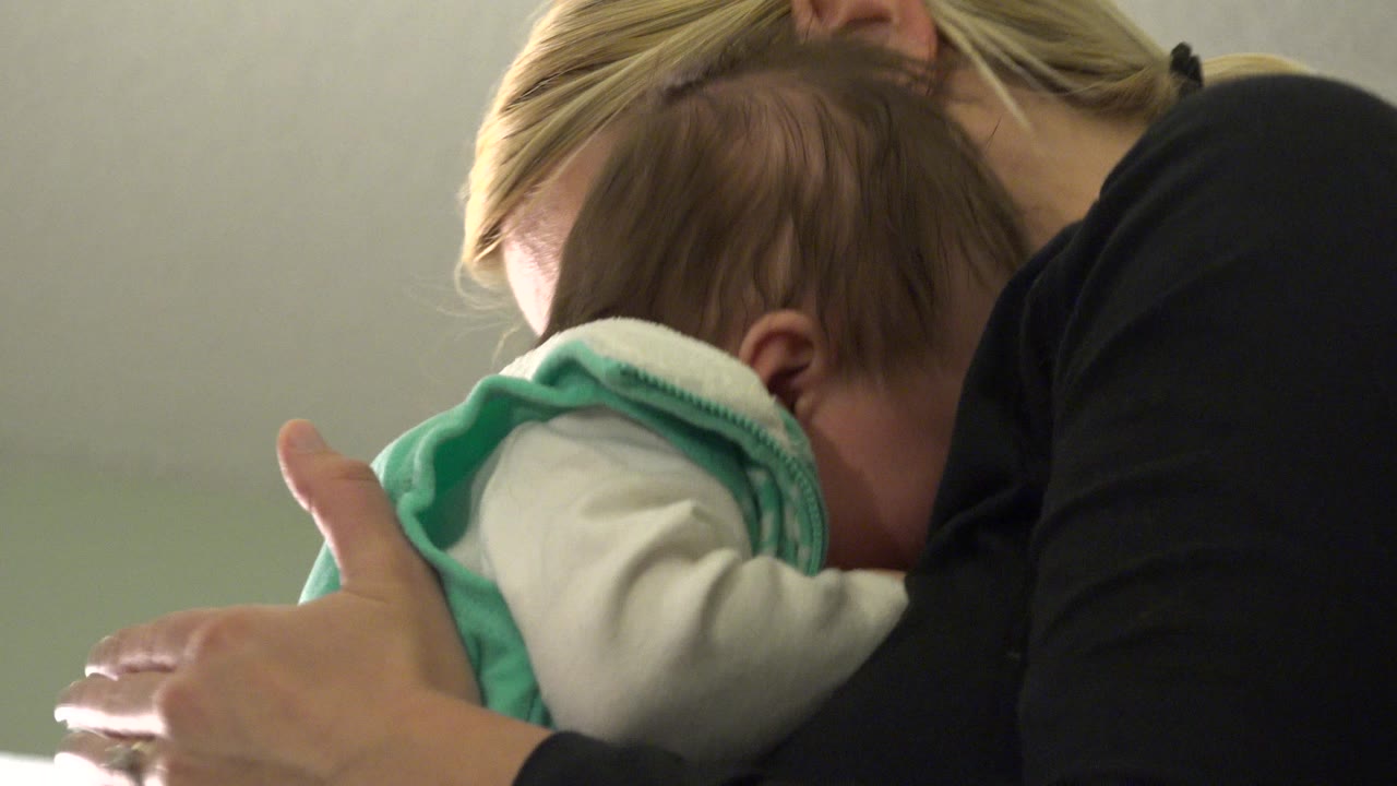 At 0:00, a blonde woman in Westminster, Colorado holds a baby close to her shoulder beside a plain pale wall.