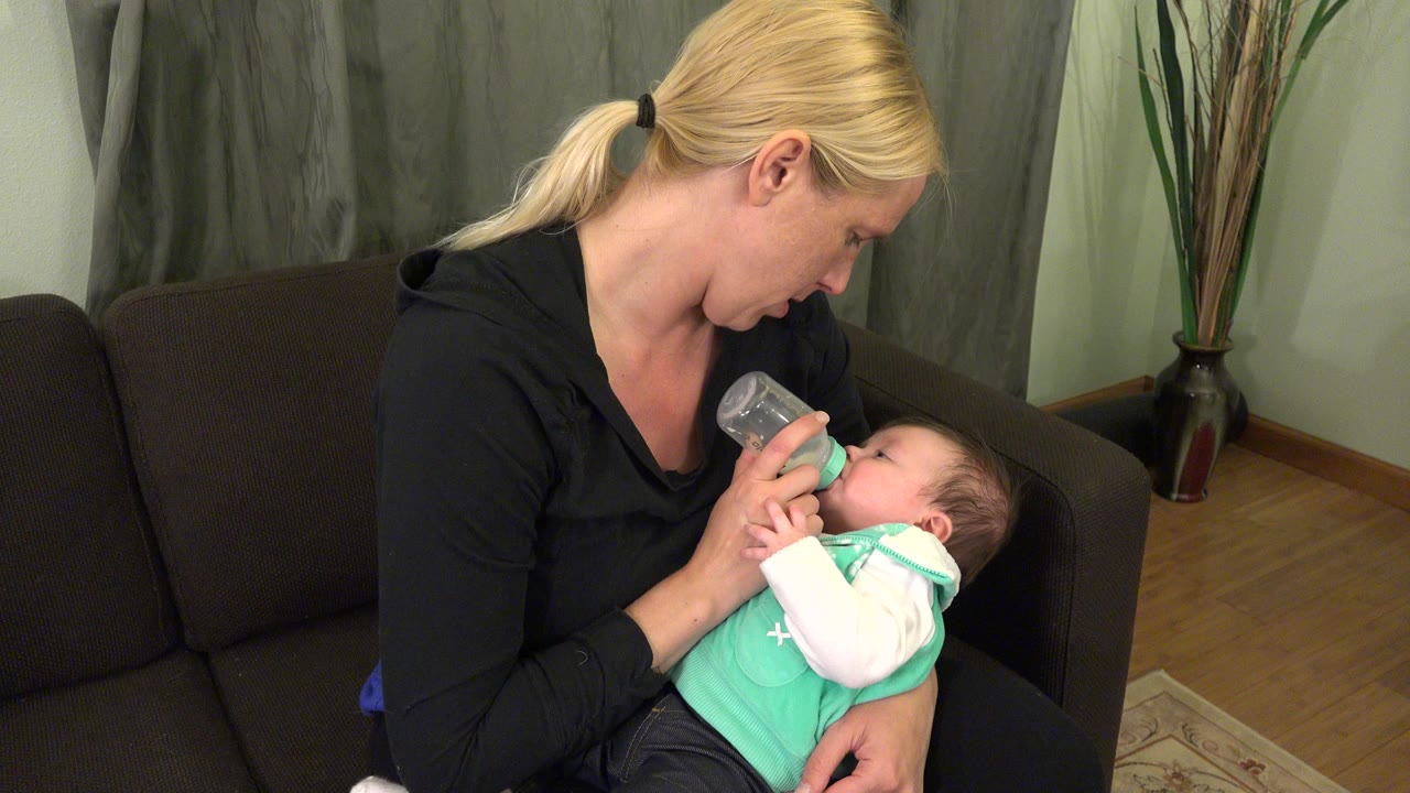 At 0:00, a blonde woman in Westminster, Colorado sits on a dark couch and holds an infant close while giving the baby a clear bottle.