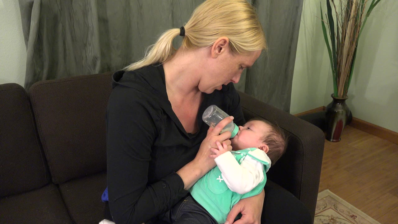 At 0:01, the woman looks down toward the infant as the bottle rests at the baby's mouth, with the couch arm and tall vase visible behind them in Westminster, Colorado.
