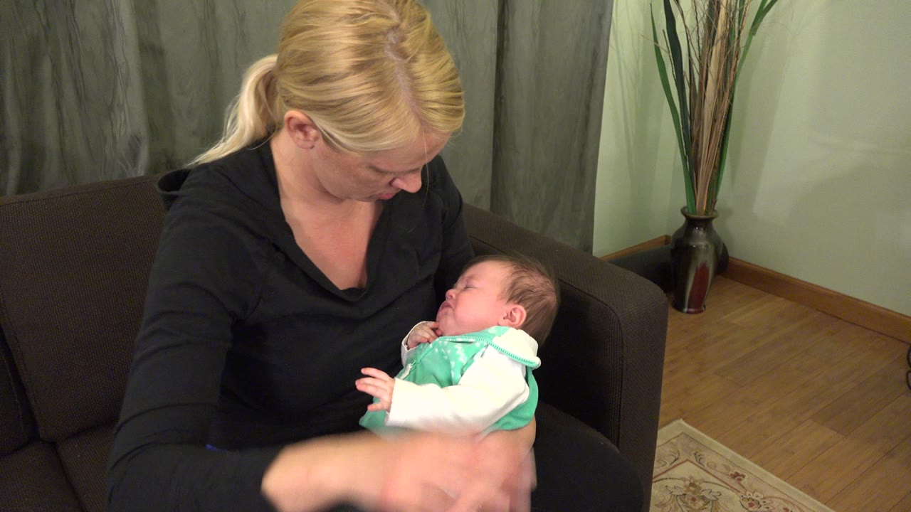 One second in, the woman keeps her gaze on the baby as the infant lies on her lap, with the sofa arm, wood floor, and decorative vase visible behind them.