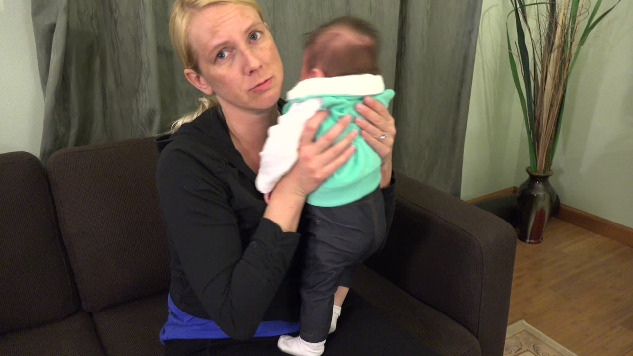 At 0:00 in Westminster, Colorado, a blonde woman sits on a dark couch holding a baby upright against her shoulder while looking toward the camera.