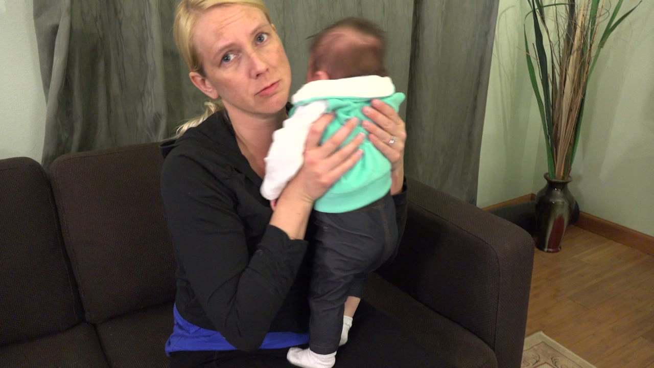 At 0:01 in Westminster, Colorado, the woman remains seated with the baby upright near her shoulder, with the couch arm and tall floor vase visible behind them.