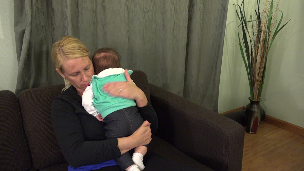 At the start of the Westminster, Colorado clip, a blonde mother sits on a dark couch with a baby held against her shoulder near green-gray curtains.