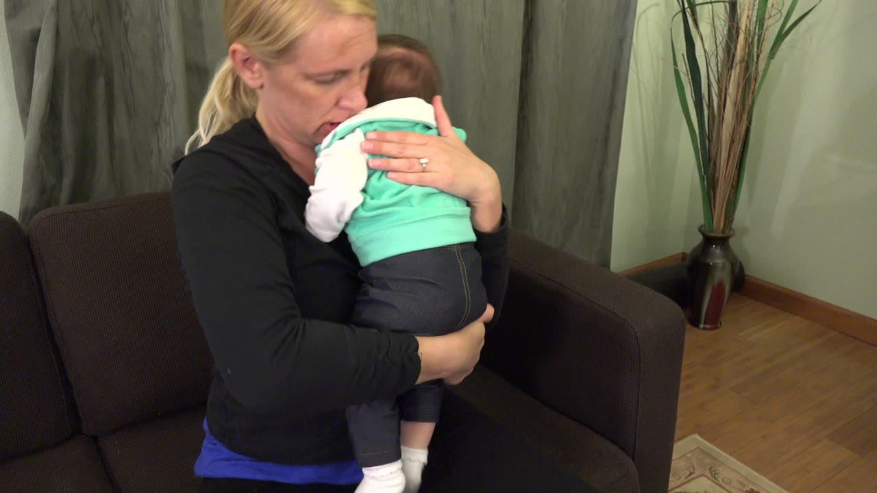 In Westminster, Colorado, a blonde mother seated beside a dark couch holds an infant in turquoise clothing close to her chest at the start of the clip.