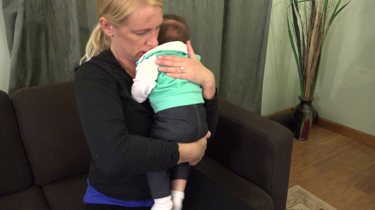 In Westminster, Colorado, the mother looks down toward the infant while one hand supports the baby's back and the couch and tall vase remain visible behind them.