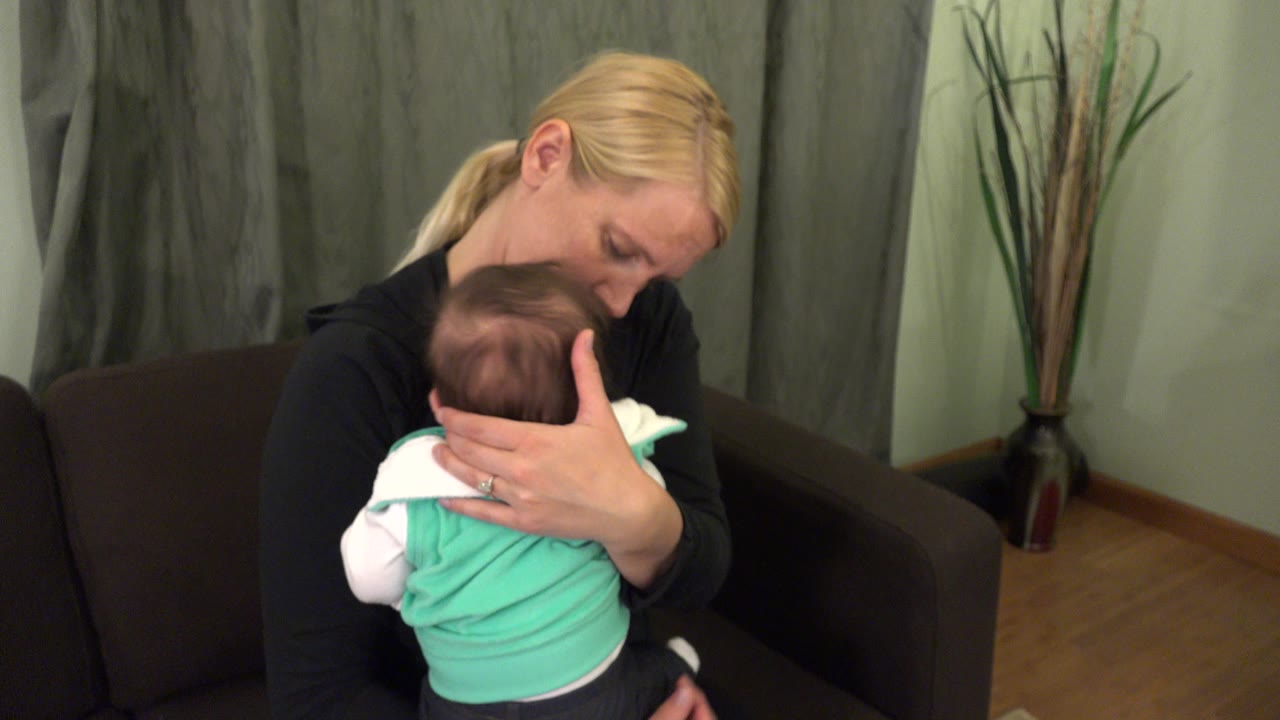In Westminster, Colorado, the mother keeps one hand behind the infant's head as she looks down from the couch, with gray curtains and a tall vase in the background.