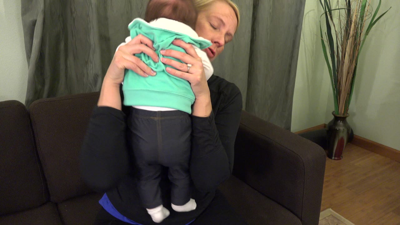 In Westminster, Colorado, a blonde mother holds a baby close against her shoulder beside a brown couch, green curtains, and a tall vase with reeds.