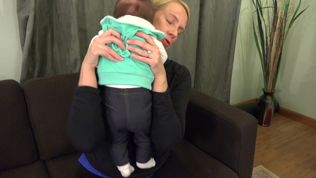 In Westminster, Colorado, the mother continues holding the baby upright with the child's back to camera and her face partly visible near the green curtains.