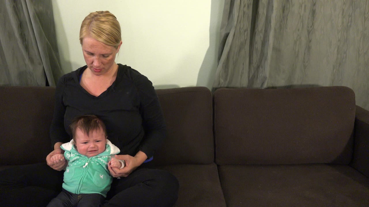At 0:00 in Westminster, Colorado, a blonde woman in a black top supports a crying infant in a mint green vest on a dark couch.