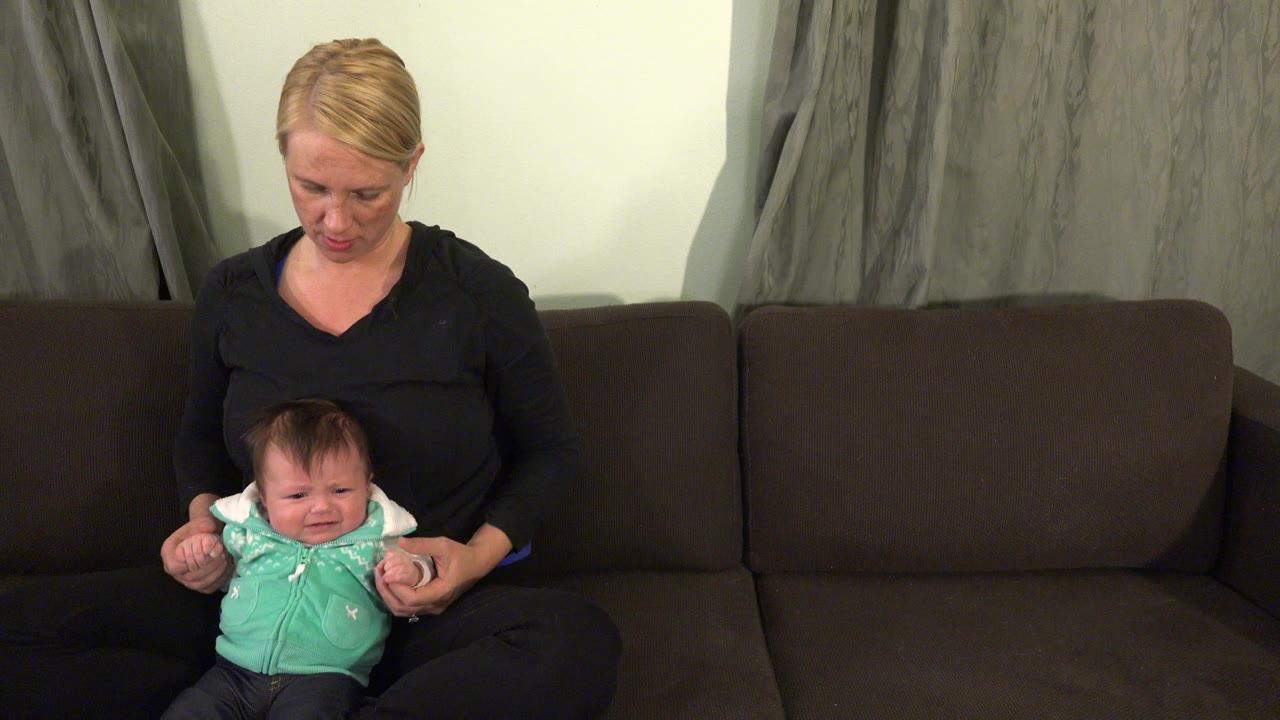 At 0:01 in Westminster, Colorado, the woman looks down while holding the infant’s hands as the baby cries on the couch.