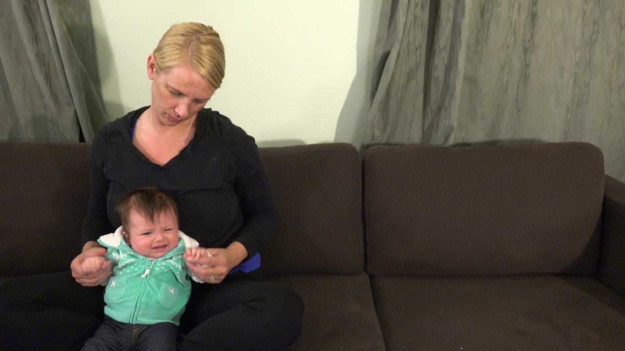 In Westminster, Colorado, a blonde mother sits on a brown couch and holds a crying baby in a mint green hoodie near gray curtains.