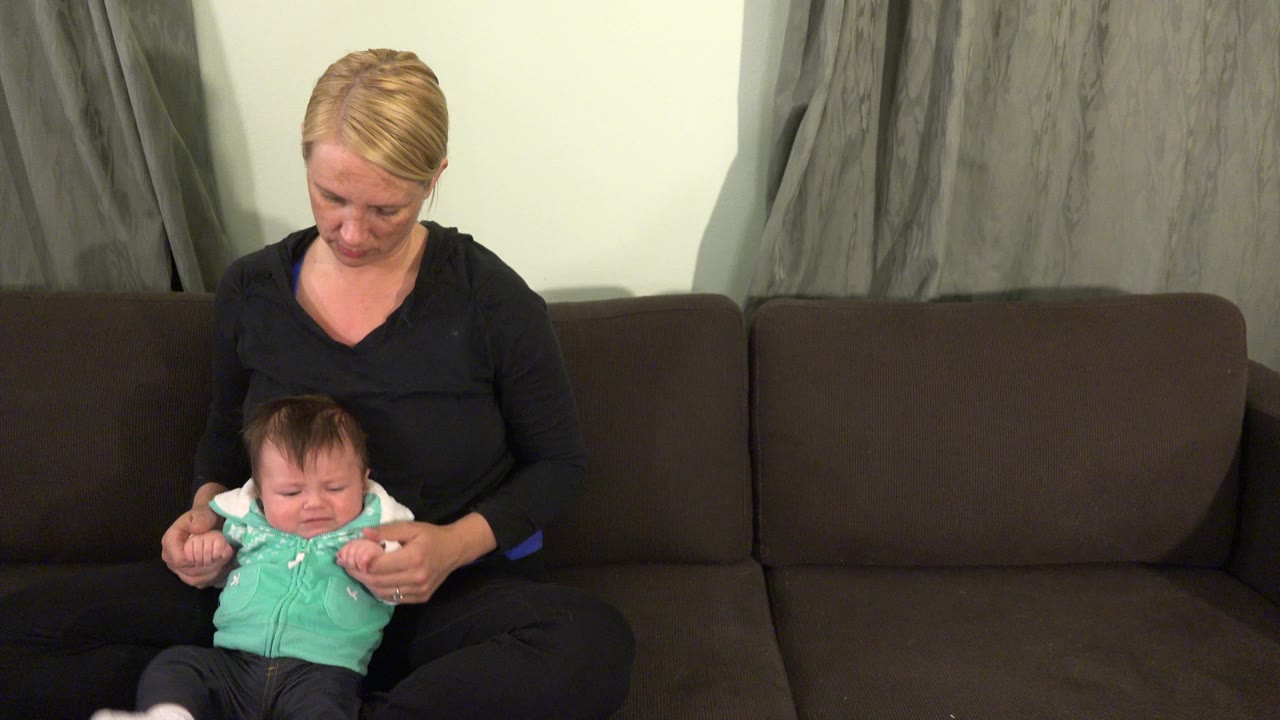 At 0:00 in Westminster, Colorado, a blonde woman in a black top looks down while holding a baby in a mint green vest on a dark couch.