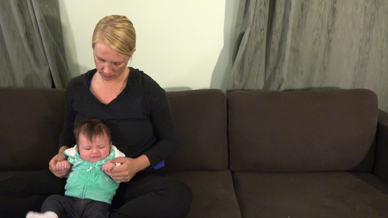At 0:01 in Westminster, Colorado, the woman keeps both hands near the baby's arms as the baby sits upright on the couch in front of gray curtains.