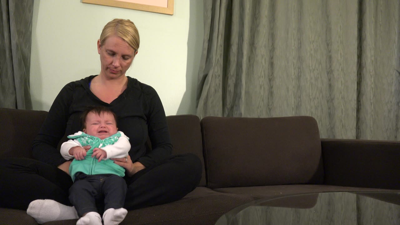 The woman looks down toward the baby as the baby sits upright on her lap, with gray curtains and dark sofa cushions behind them in the Westminster home.