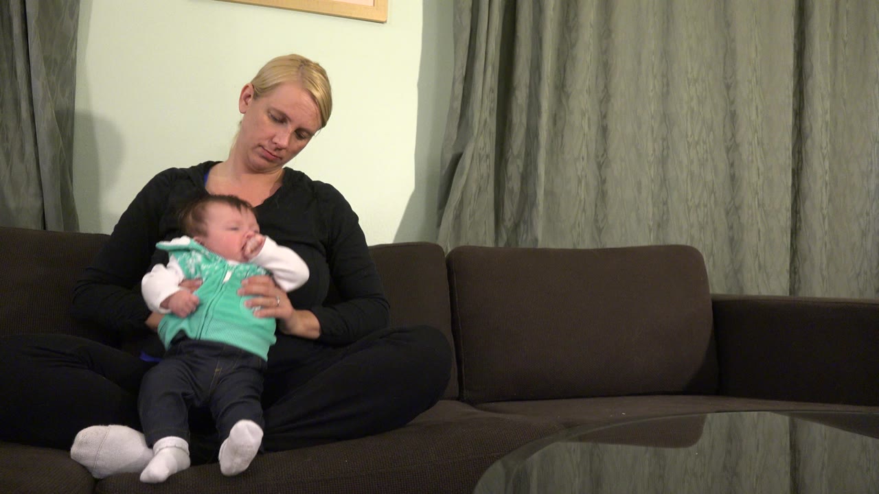 In Westminster, Colorado, a blonde woman sits cross-legged on a brown sofa while holding a baby in a mint green vest near gray curtains.