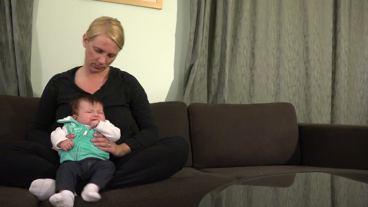The mother in Westminster, Colorado looks down toward the crying baby, whose hand is near the mouth while both remain seated on the couch.