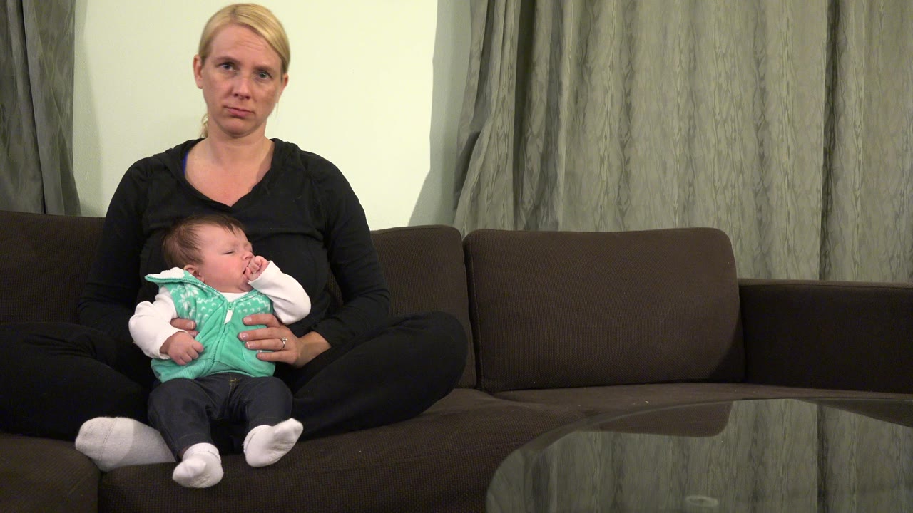 Woman holding a baby on a dark sofa in a living room.