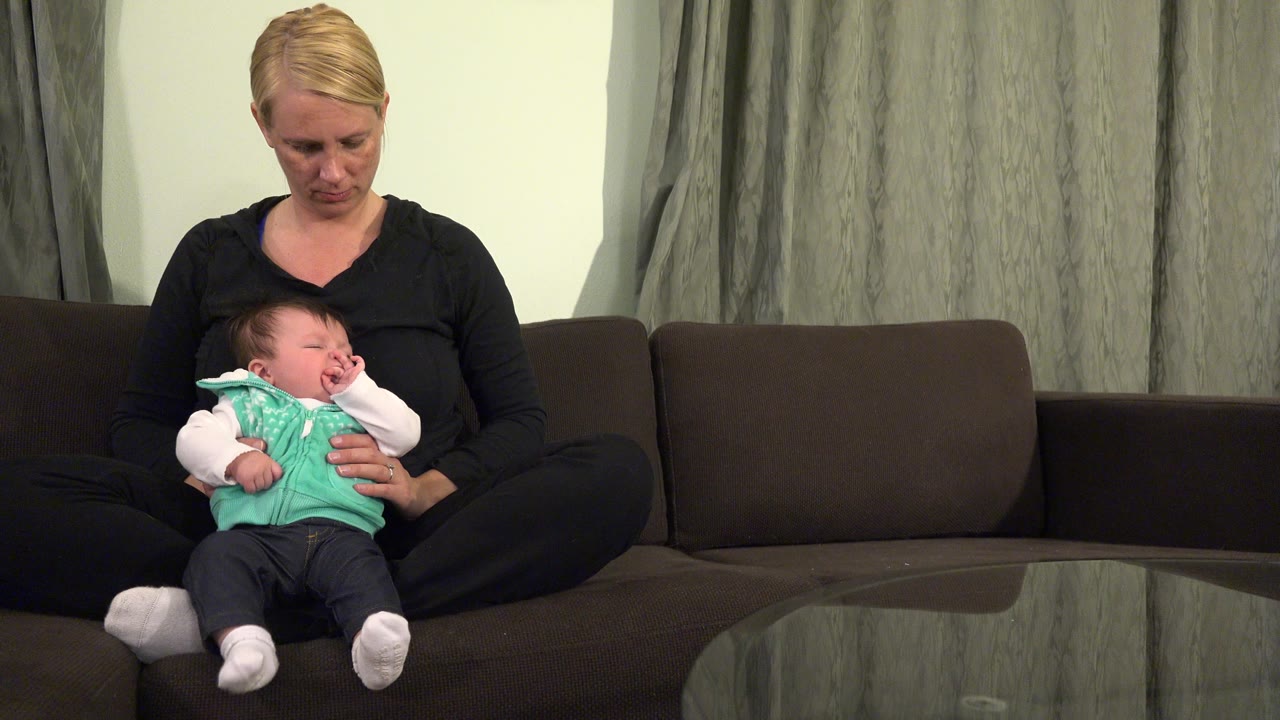 In Westminster, Colorado, a blonde woman looks down while holding a baby in a green vest on a dark couch beside gray curtains and a glass table.