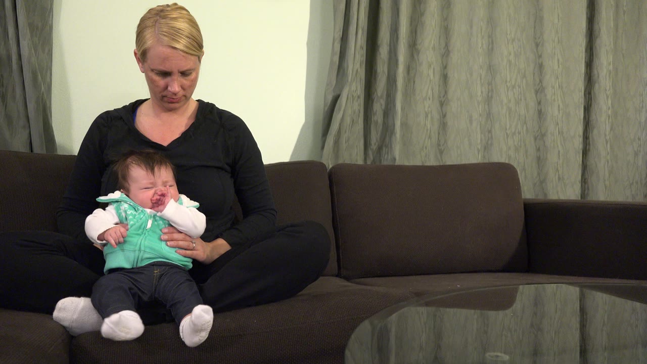 In Westminster, Colorado, the woman looks down at the baby in her lap while seated cross-legged on the sofa, with gray curtains behind them.