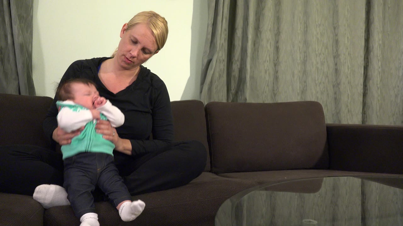 In Westminster, Colorado, the woman looks down at the crying baby she is supporting on the sofa, with the baby's white sleeves and turquoise shirt visible against the dark couch.