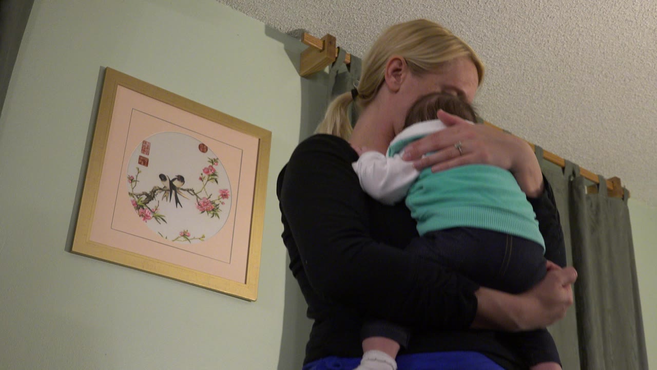 Mother holding a sleeping baby indoors beside framed wall art.
