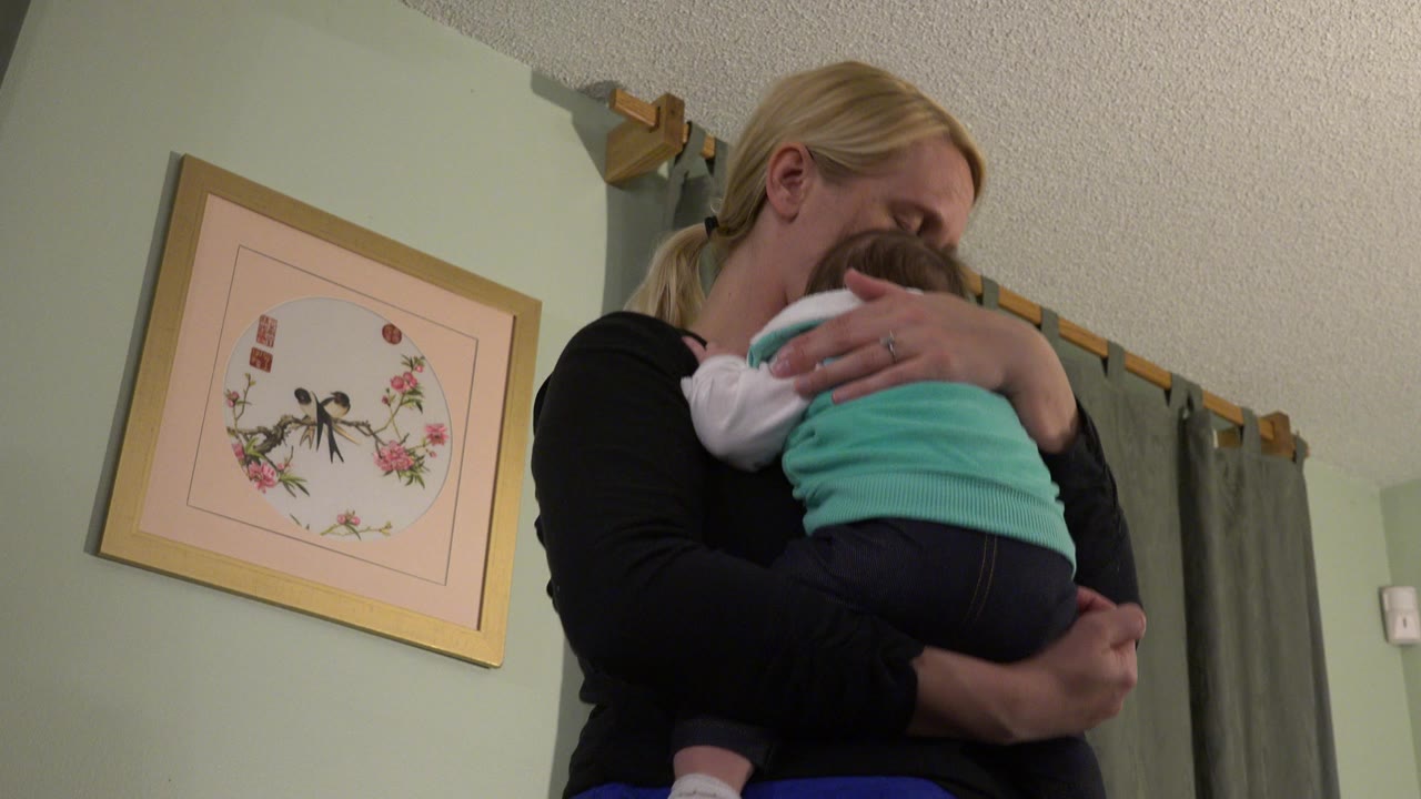 Woman holding a small baby indoors beside framed wall art.