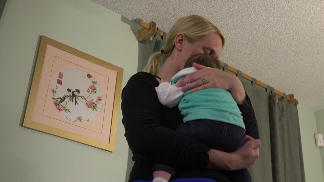 At 0:01 in Westminster, Colorado, the woman keeps the baby against her chest as her face lowers toward the child's head, with the framed artwork still visible on the left wall.