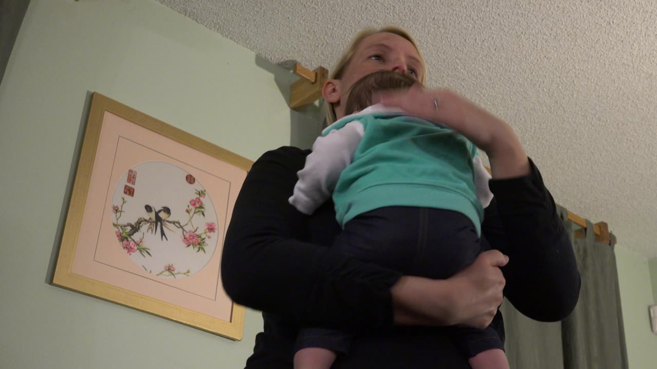 At 0:01 in Westminster, Colorado, the baby rests higher on the mother's shoulder while her hand supports the child, with the framed wall art still visible at left.