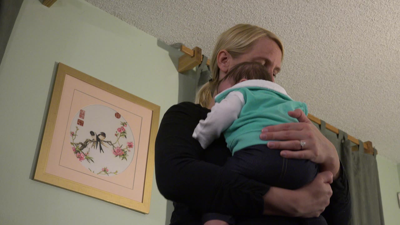 Mother holding a baby indoors beside framed wall art.