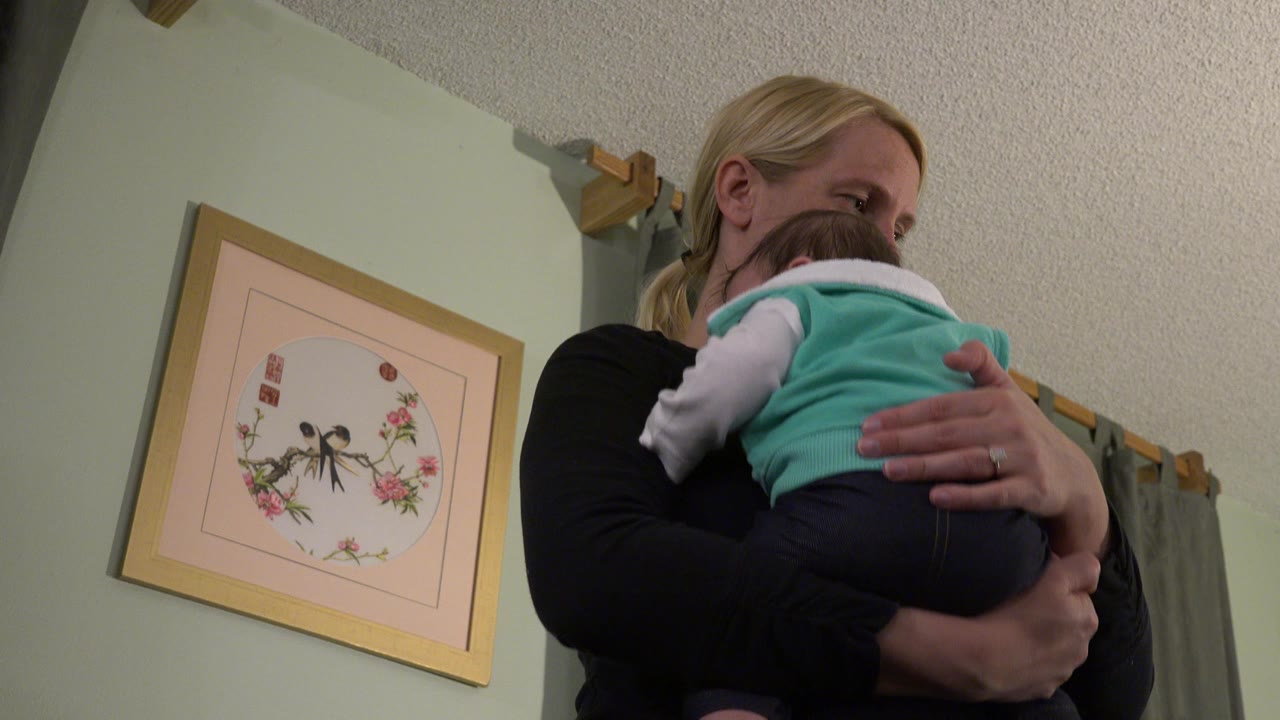 At 0:01 in Westminster, Colorado, the woman keeps the baby against her chest while her face is partly turned downward and the framed artwork remains clear on the left wall.