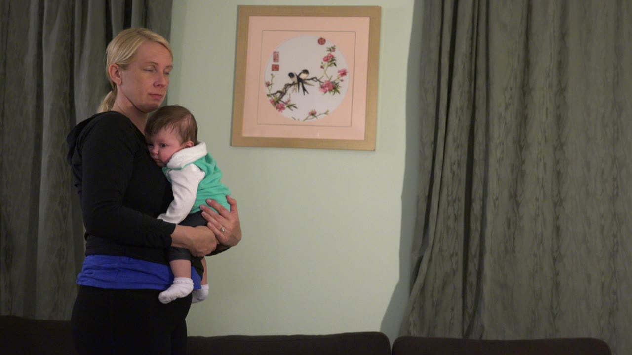 Mother holding a baby indoors beside gray curtains.