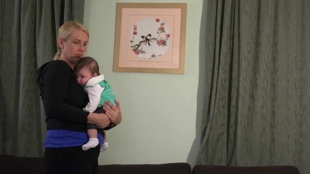 At 0:00 in Westminster, Colorado, a blonde woman holds a baby against her chest beside gray curtains and a framed picture on a pale green wall.