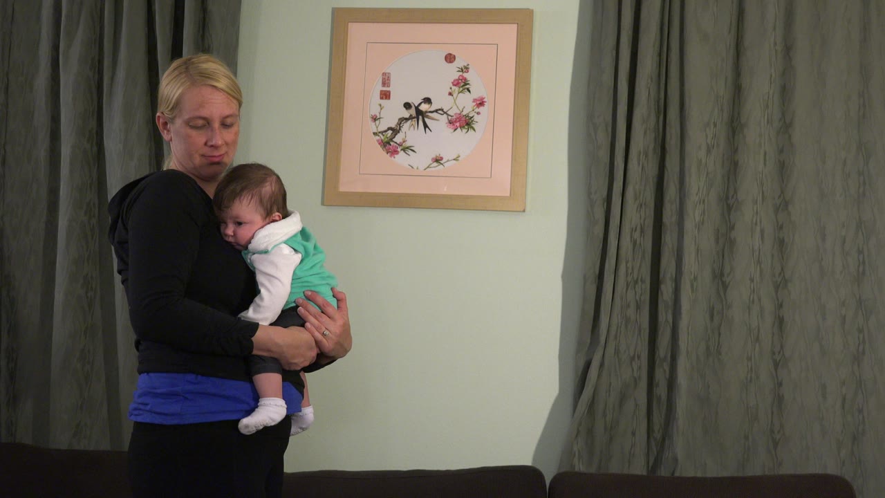 Blonde woman holding a sleeping baby indoors beside curtains.
