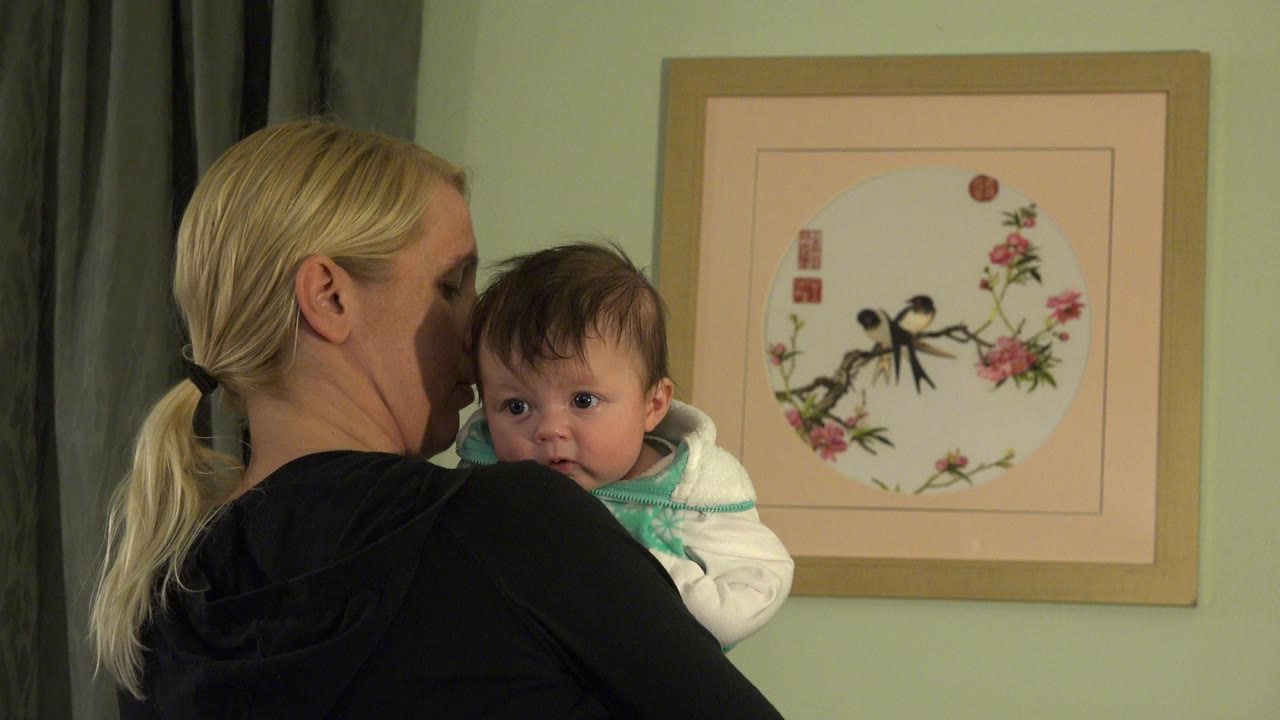 Blonde woman holding a baby beside framed bird artwork.