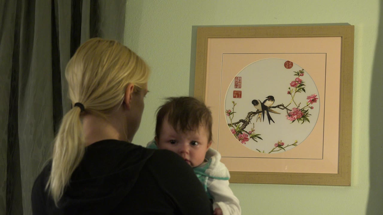 Blonde woman holding a baby beside framed bird artwork.