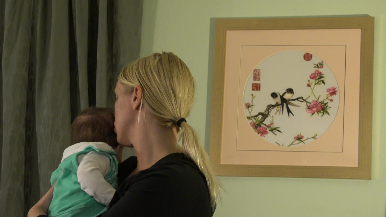 Blonde woman holding a baby beside curtains and framed wall art.