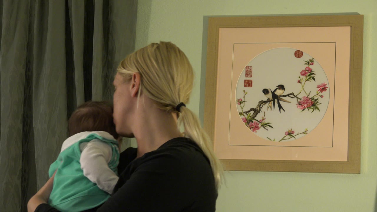 Mother holding a baby beside gray curtains and framed wall art.