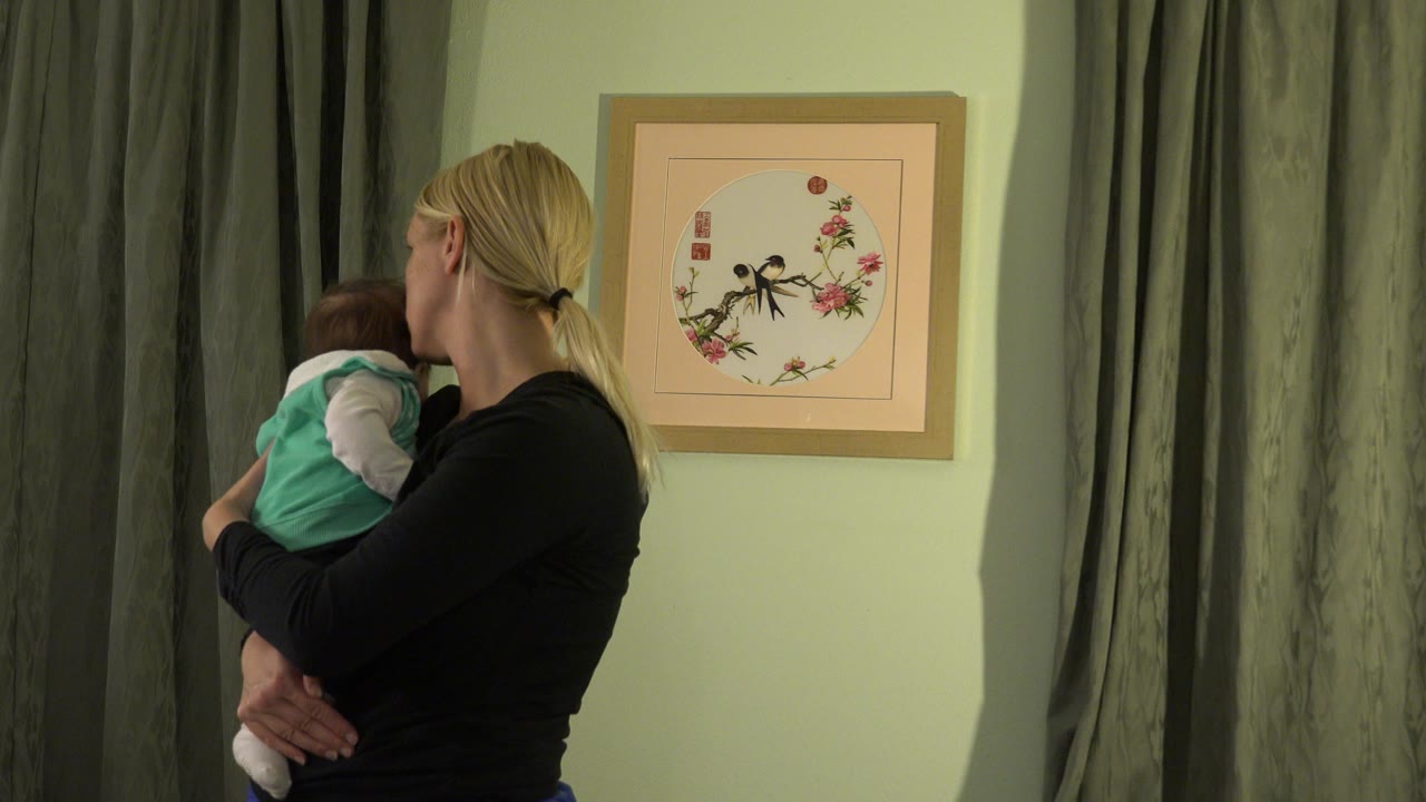 Mother holding a baby beside curtains and framed wall art.