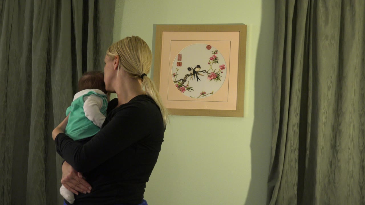 In Westminster, Colorado, the mother keeps the baby against her shoulder while standing in front of green curtains and a framed bird-and-flower artwork.