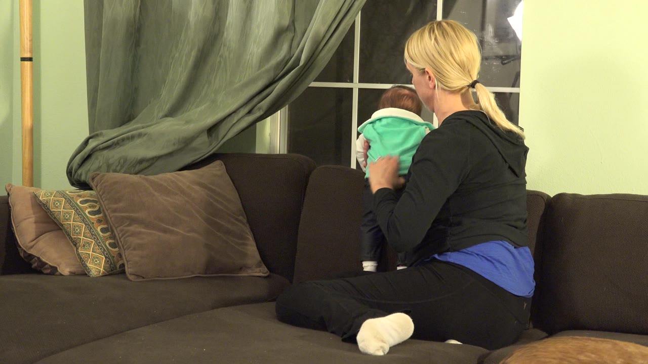 Blonde woman and baby seated on a sofa beside a window.