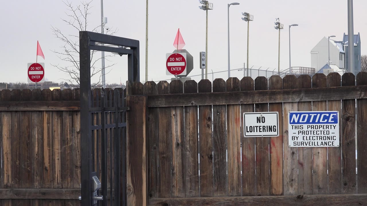 Opening frame of a dark wooden fence in Westminster, Colorado, with a black metal gate at left, red do-not-enter signs beyond it, cloudy daylight, and no visible people.