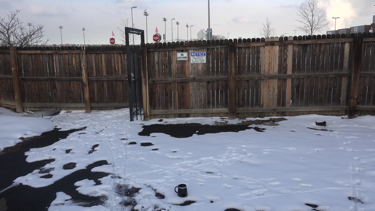 At 0:01 in Westminster, Colorado, the fence fills the midground with warning signs near center, snow patches below, poles above the fence, and no visible people.