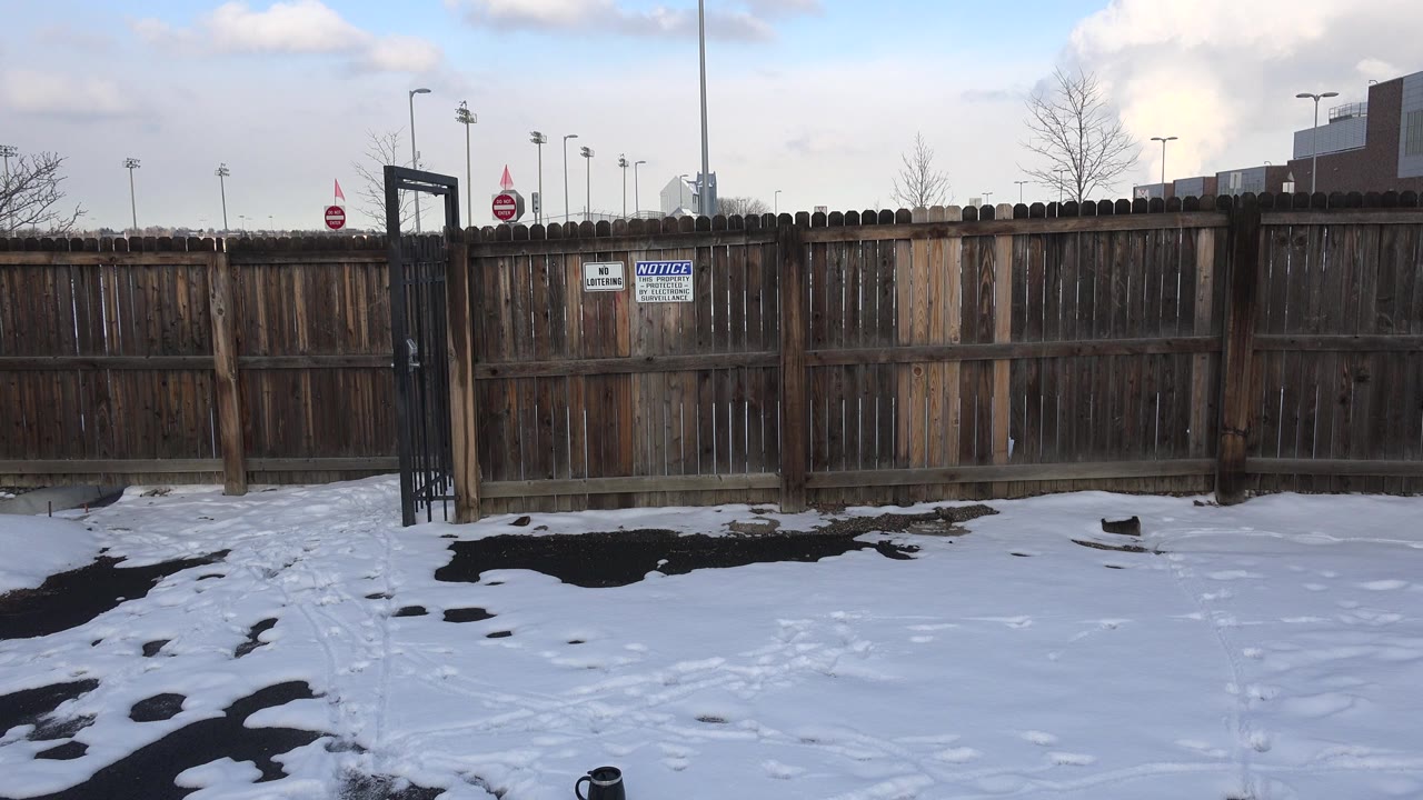 Opening frame in Westminster, Colorado showing patchy snow, dark pavement, a wooden plank fence, a black metal gate, posted signs, distant light poles, and no visible people.