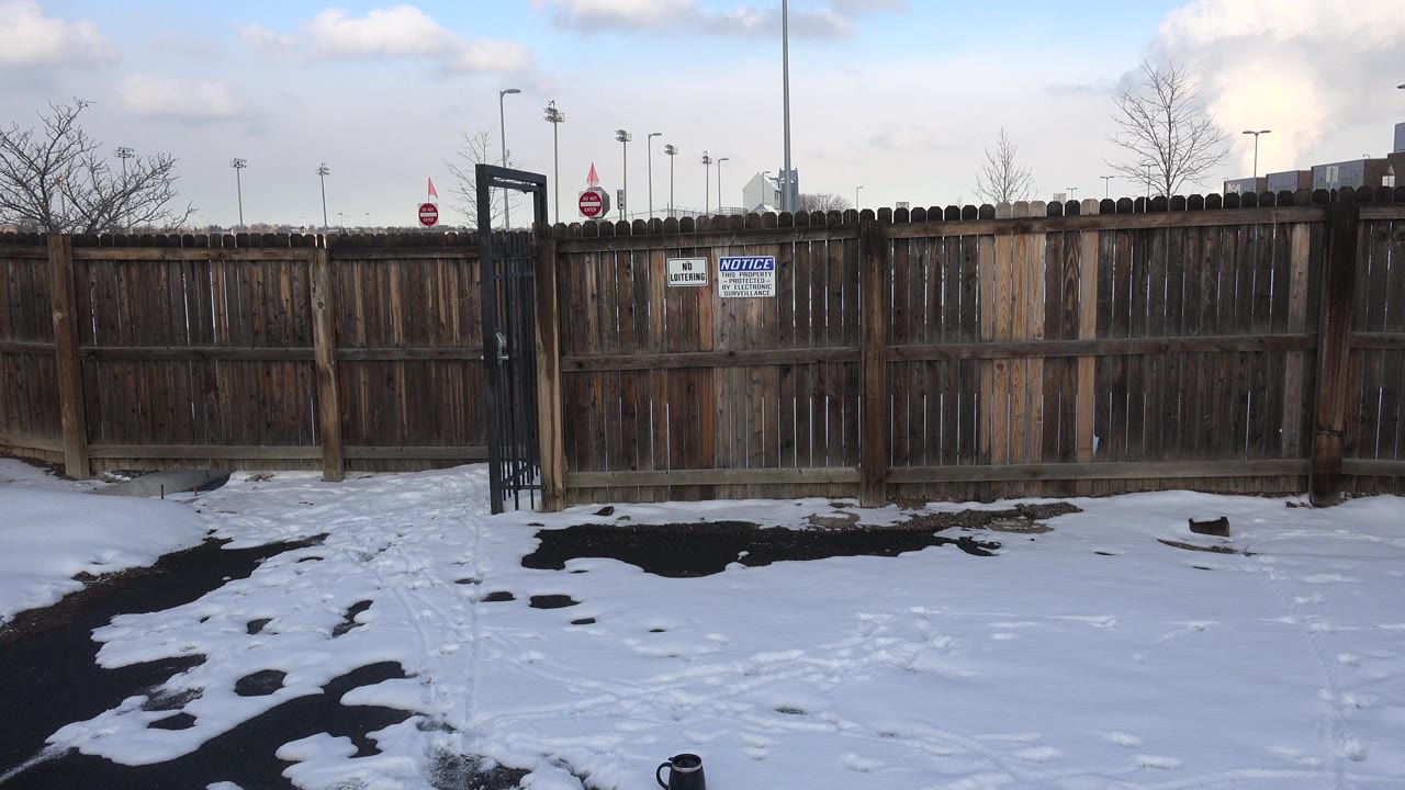 Opening frame in Westminster, Colorado shows snow patches and dark pavement leading to a weathered wooden fence, a black metal gate, posted signs, bare trees, and no visible people.