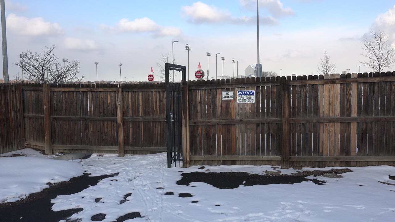 Opening frame of a brown wooden fence in Westminster, Colorado with snow patches in front, light poles behind the fence, and no visible people.