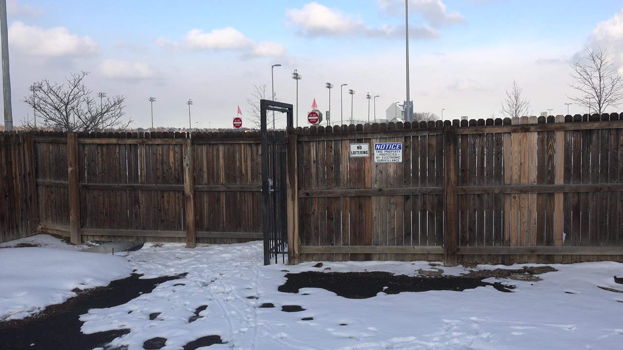 Weathered brown fence in Westminster, Colorado with a black gate near center, snow along the pavement, warning signs on the boards, and no visible people.